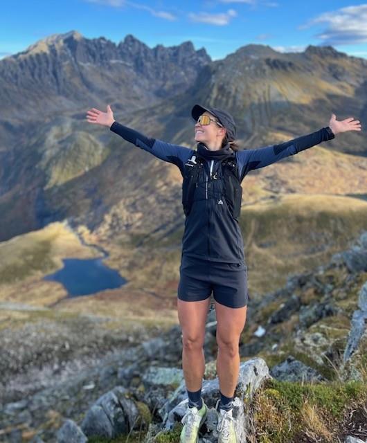 kvinne ikledd turklær, solbriller og caps poserer med armene utstrakt på en fjellsti, i bakgrunnen ruger høye fjelltopper