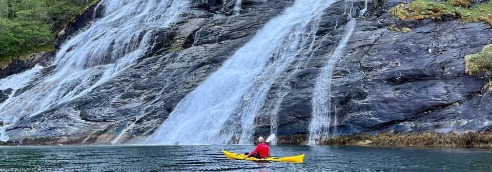 en person padler i kano på vatnet, i bakgrunnen er berg og en stor foss som renner ut i vannet