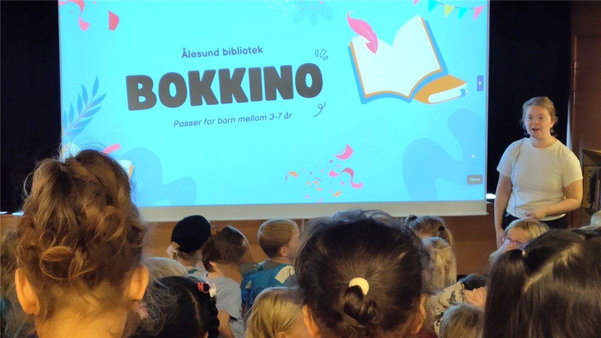 Vi ser et lerret der det står BOKKINO Ålesund bibliotek passer for barn mellom 3 - 7 år. Foran lerretet ser vi bakhodene på en flokk barn i førskolealder. Ved siden av står en bibliotekar og leser høyt.