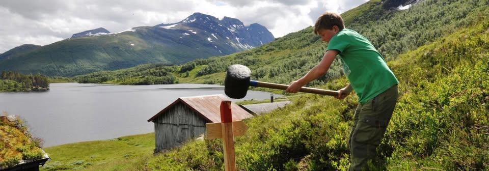 en ung gutt slår en påle i marken med ei slegge. i bakgrunnen er et vann og en gård og fjell.